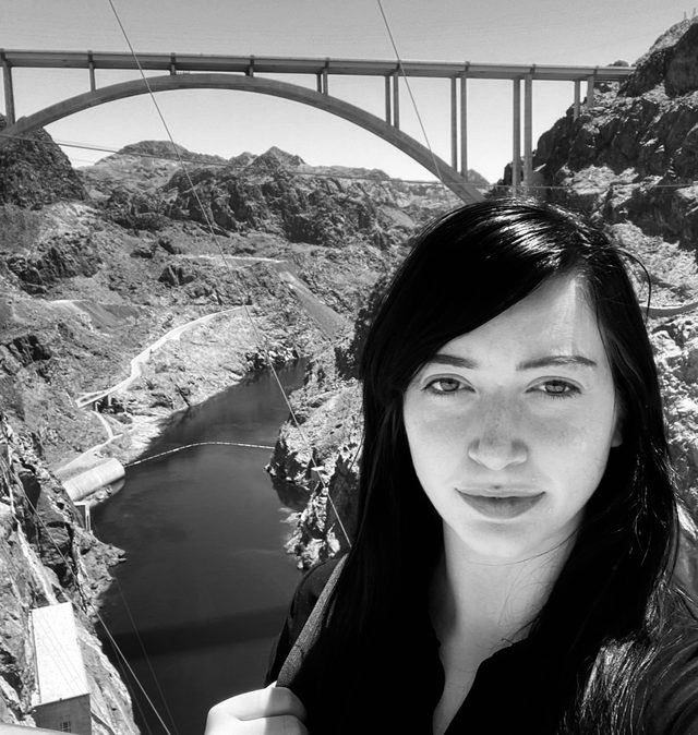 Iris taking a selfie above a canyon, with a concrete arch bridge behind her
